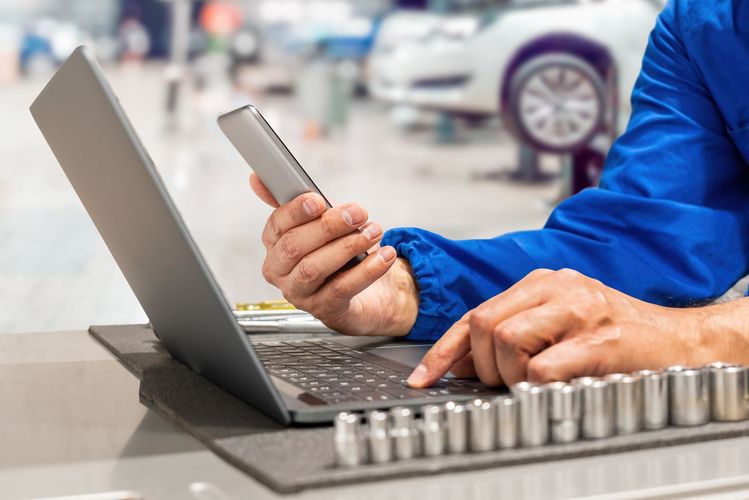 Un mécanicien en combinaison bleue utilise un ordinateur portable et un téléphone, avec des outils et une voiture en arrière-plan.