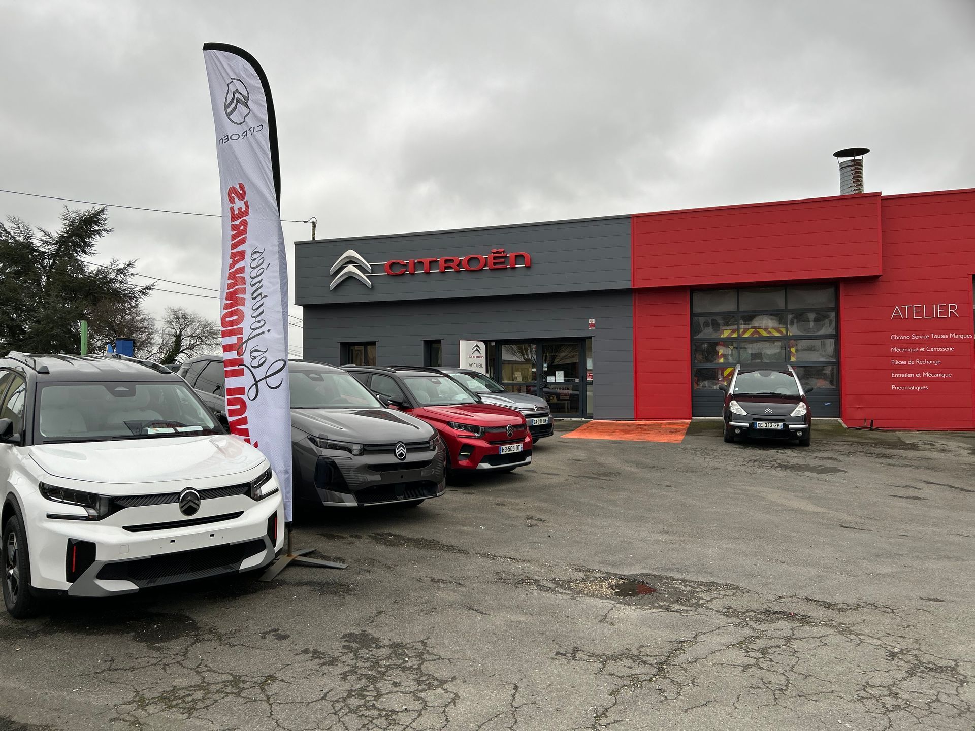 Concessionnaire Citroën avec voitures garées à l'extérieur, bâtiment rouge et gris, journée nuageuse.
