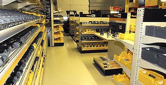 Allée d'entrepôt avec des étagères métalliques remplies de cartons et de pièces détachées sous un éclairage fluorescent intense