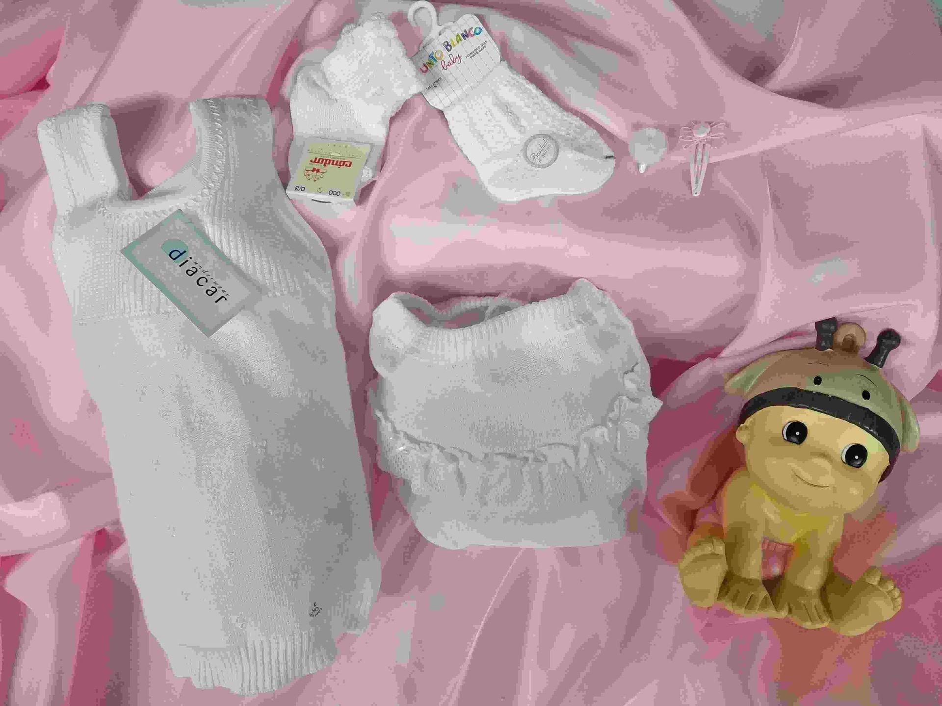 Una muñeca está sentada al lado de una pila de ropa de bebé.