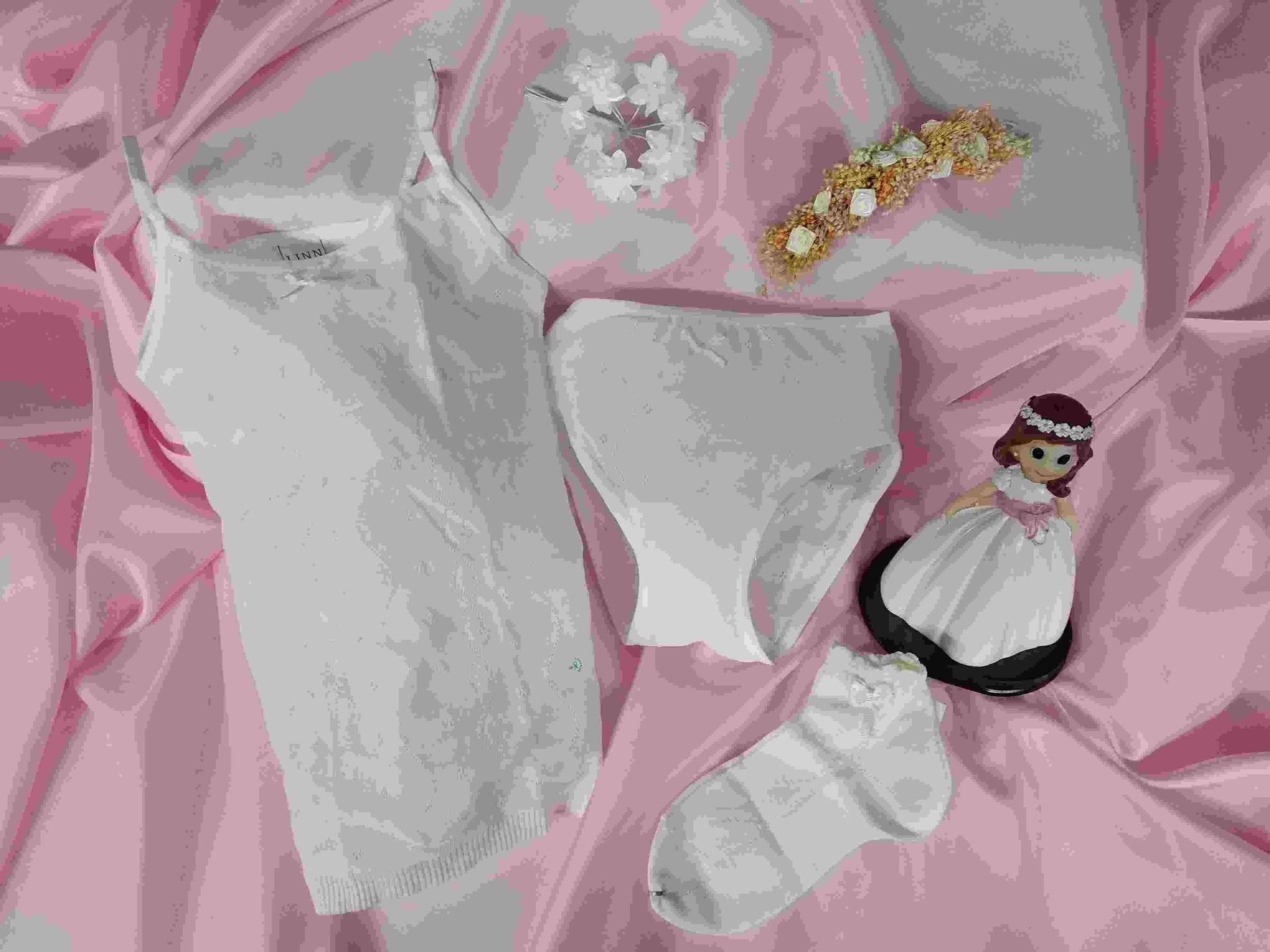 Una muñeca con un vestido de novia está sentada sobre una tela rosa al lado de un montón de ropa blanca.