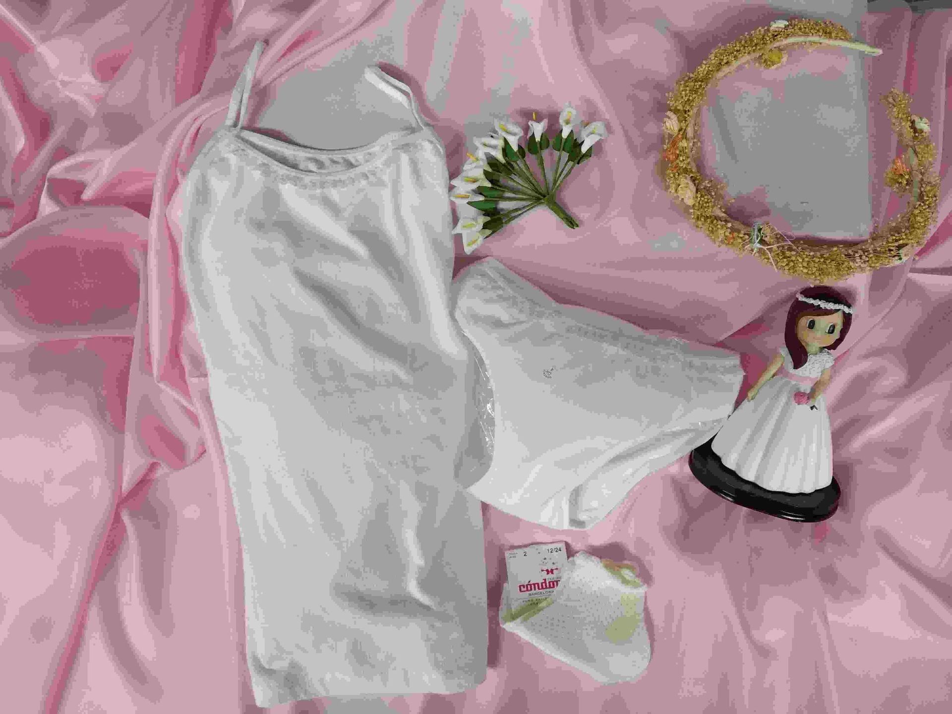 Una muñeca está sentada sobre una tela rosa al lado de un vestido y ropa interior blancos.