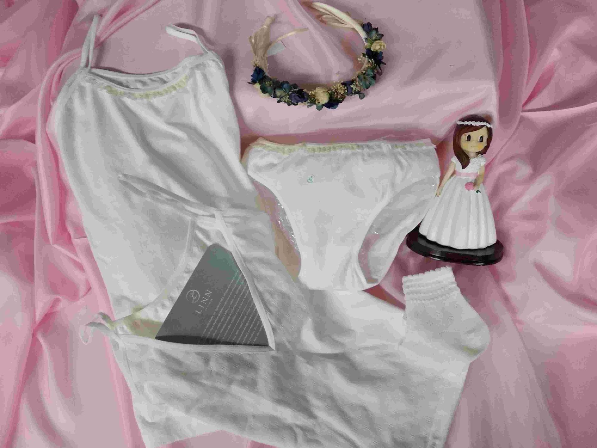 Una muñeca está sentada sobre una tela rosa junto a ropa interior blanca y una corona de flores.