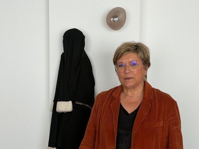 Une personne portant une veste se tient à côté d'un manteau et d'un chapeau accroché au mur, dans une pièce simple.