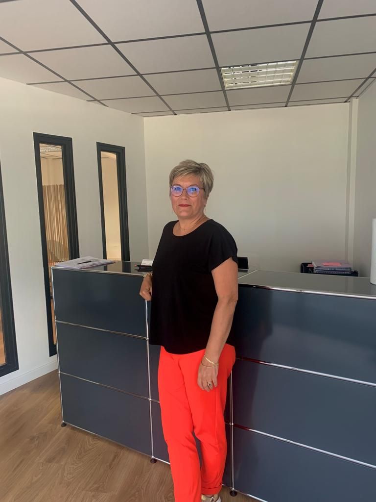 Une personne se tient derrière un comptoir d'accueil dans le hall d'un immeuble de bureaux, vêtue d'une chemise et d'un pantalon.