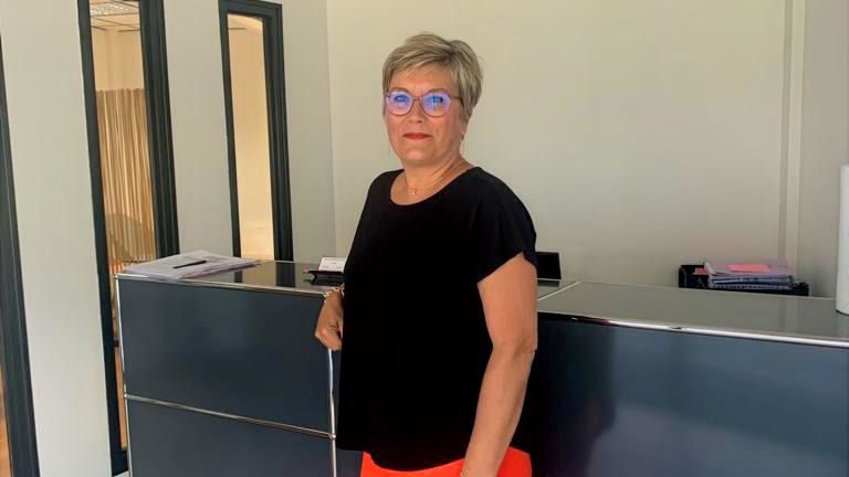 Une femme se tient debout dans un hall, près d'une réception, vêtue d'un haut et d'un pantalon.