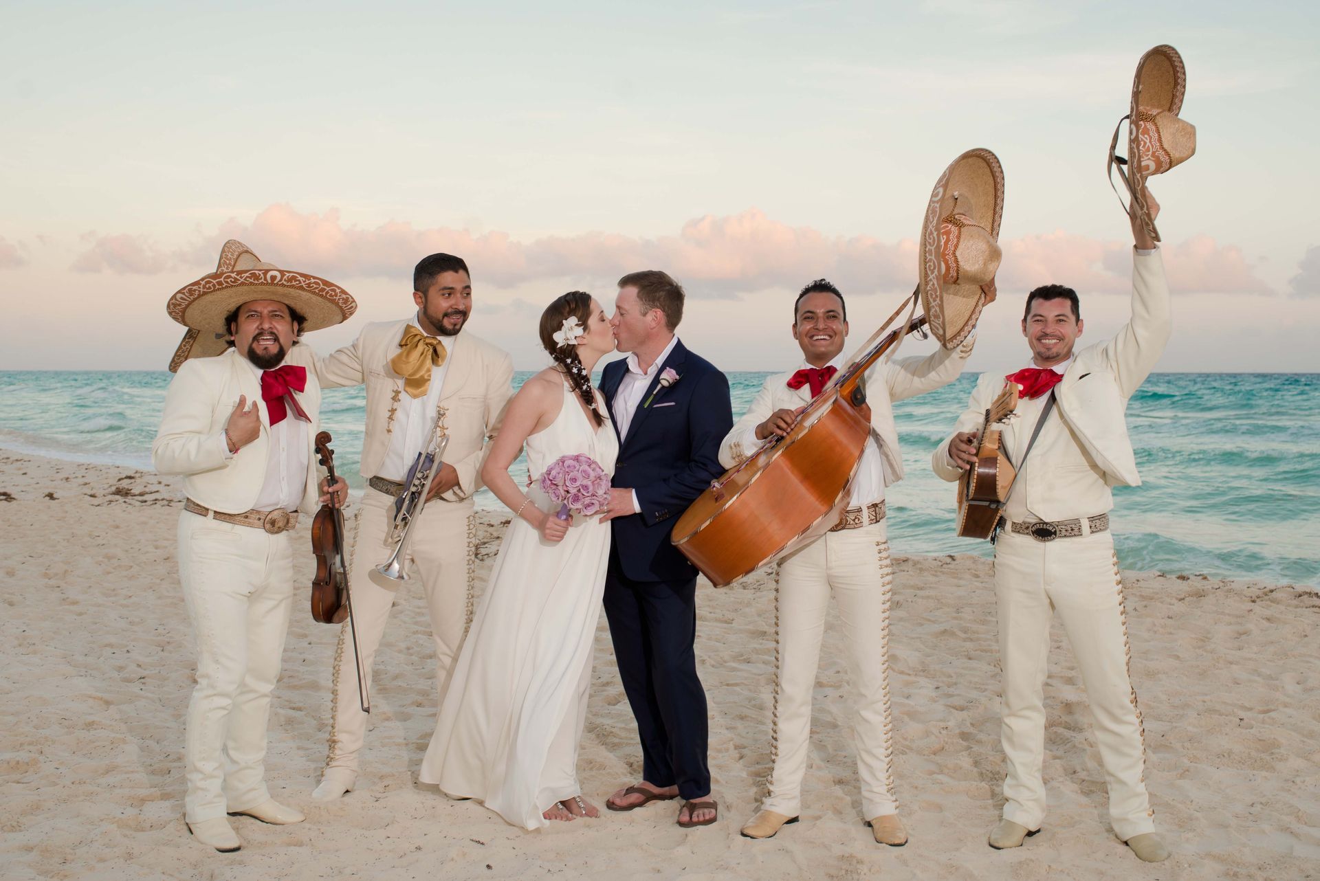 A bride and groom are kissing in front of a mariachi band on the beach.