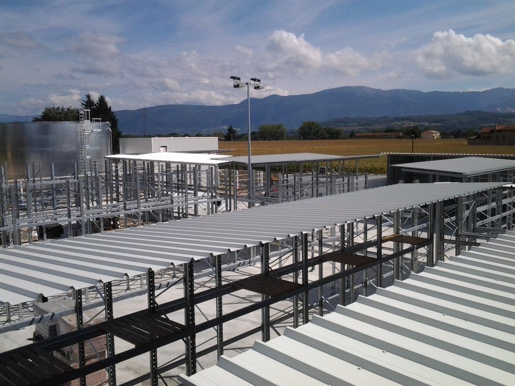 Toit industriel avec conduits, tuyaux et rambardes métalliques se détachant sur des montagnes lointaines et un ciel nuageux.