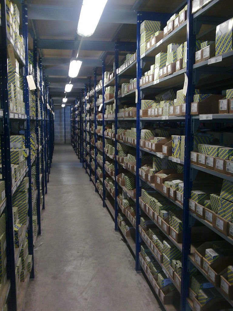 Une allée étroite d'entrepôt, bordée d'étagères métalliques remplies de cartons et de fournitures, sous des lumières fluorescentes.