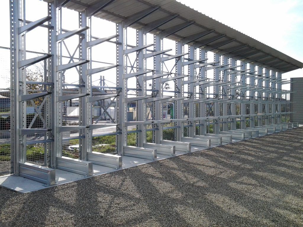 Des étagères métalliques, disposées en rangées dans une structure extérieure couverte, sur un sol en gravier.
