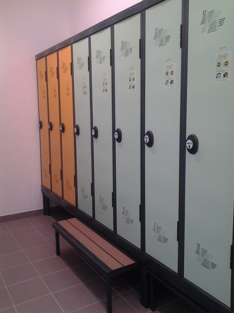 Rangée de portes de casiers orange et grises avec poignées noires dans un vestiaire carrelé, avec un banc devant.