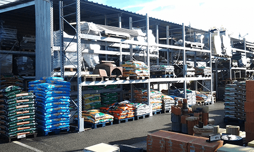 Allée d'un magasin de bricolage en plein air, avec des étagères remplies de fournitures et de palettes baignées de soleil.