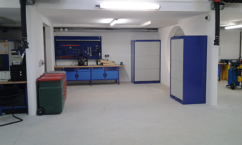 Atelier spacieux dans un garage avec des armoires bleues, un établi et une poubelle verte.