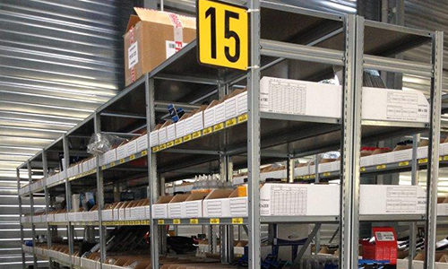 Rayonnages d'entrepôt remplis de cartons blancs, signalés par un grand panneau jaune indiquant l'allée 15