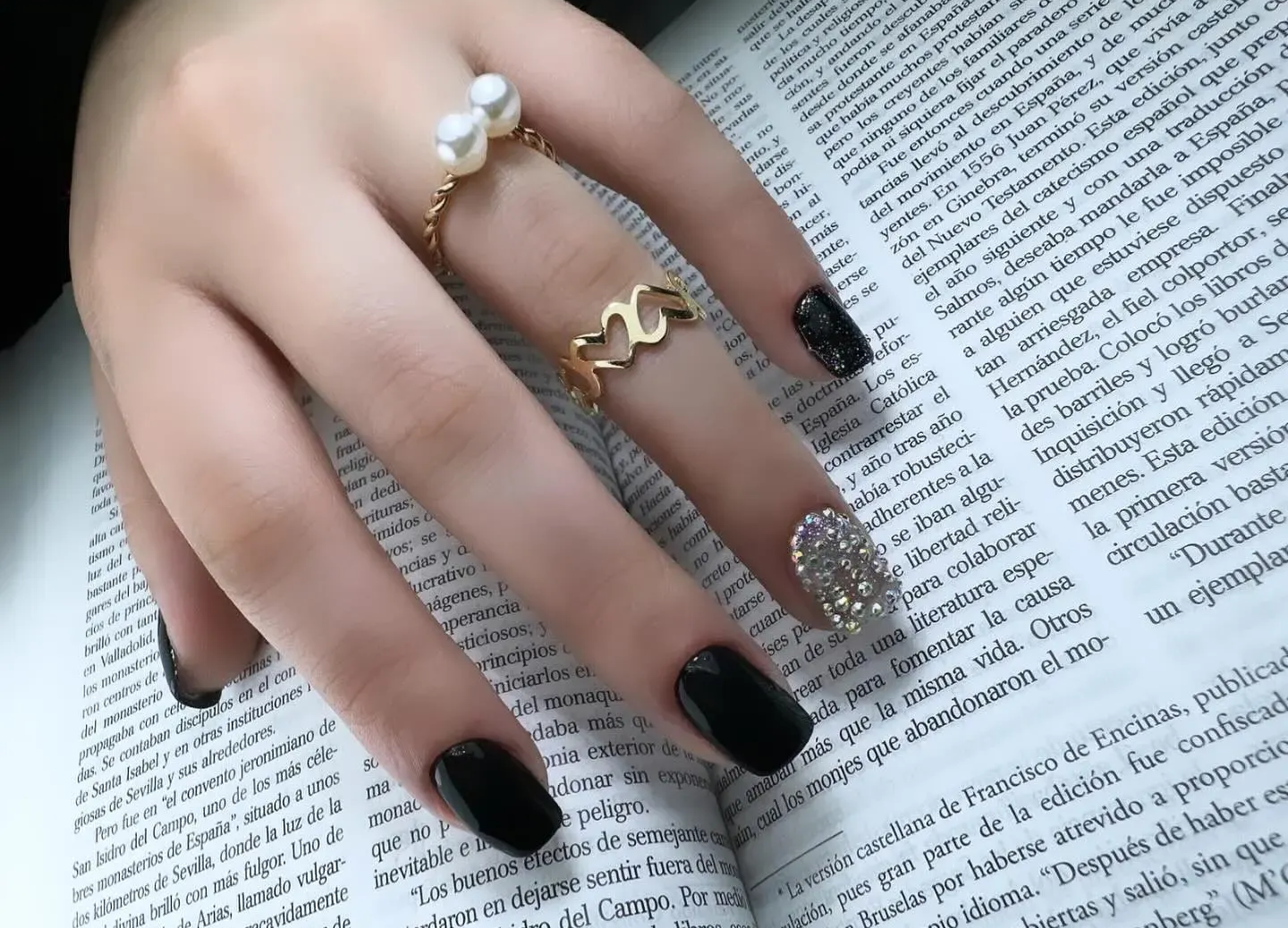 Mano con esmalte de uñas negro y anillos de oro apoyada sobre la página de un libro abierto.