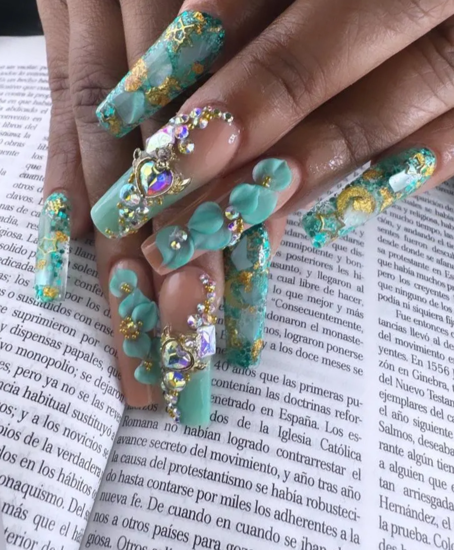 Uñas acrílicas con joyas turquesas y doradas sobre la página de un libro abierto.