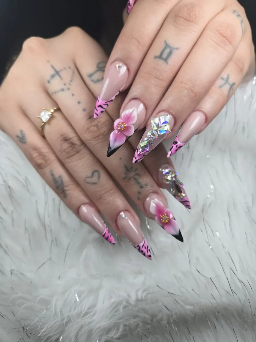 Manos con largas uñas acrílicas rosas con purpurina y tatuajes, descansando sobre una superficie de piel blanca.