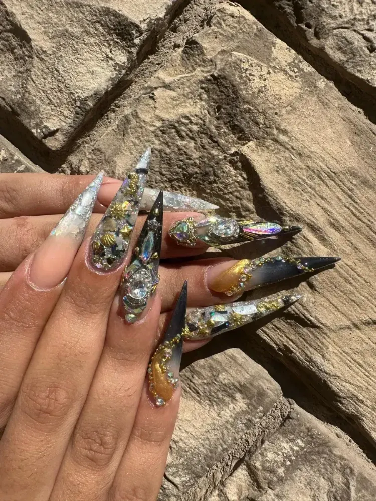 Uñas largas de aguja metálicas con purpurina y detalles de gemas sobre un fondo de piedra.