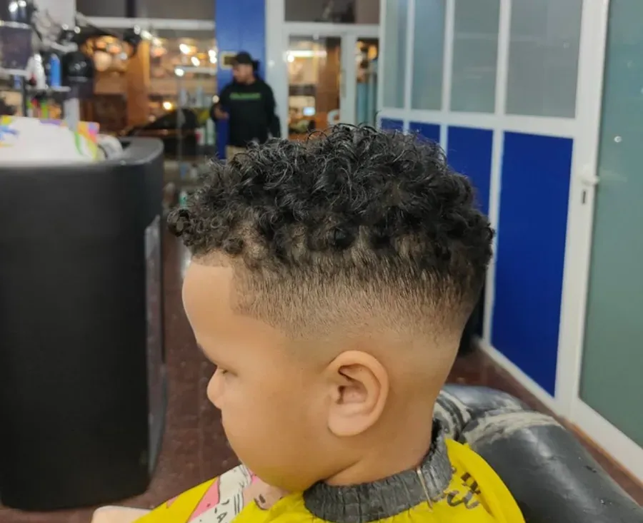Niño con corte de pelo rizado degradado sentado en una silla de barbería, de perfil.