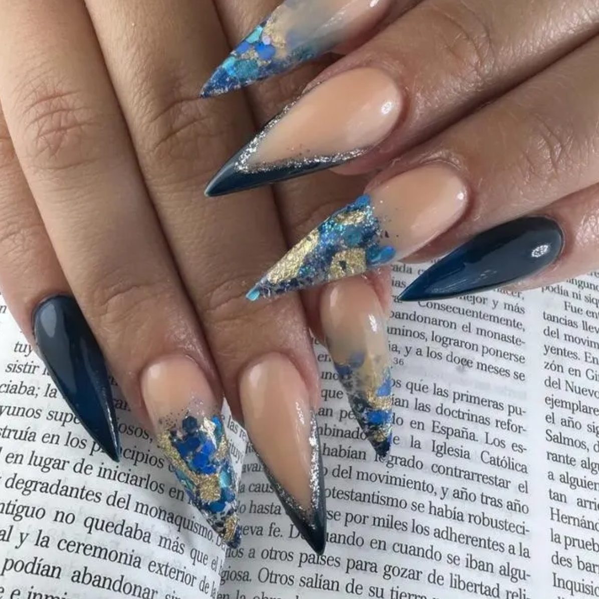 Uñas acrílicas largas de color nude y azul con forma de aguja y detalles dorados sobre una página impresa.