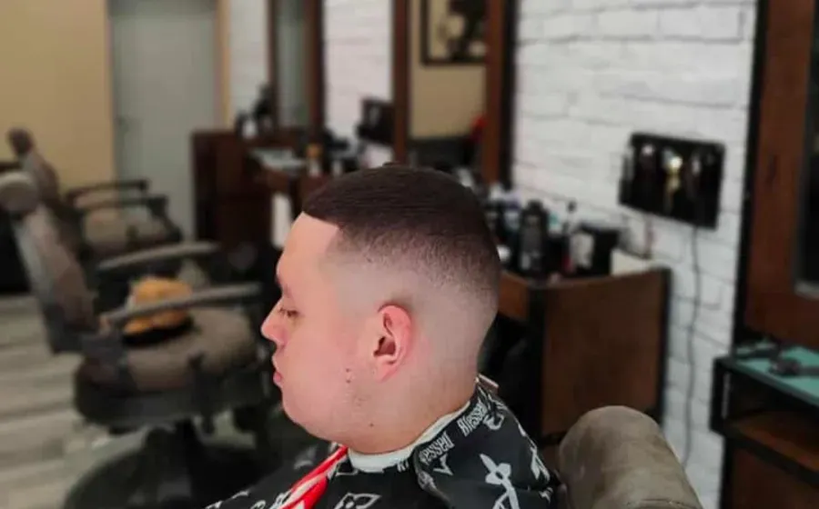 Perfil lateral de una persona sentada en una silla de barbería con el pelo recién cortado, con sillas de peluquería y espejos detrás.