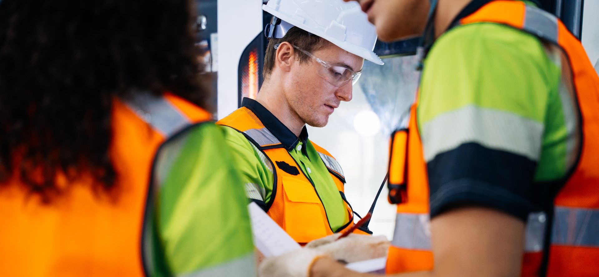 Trois ouvriers, vêtus de gilets orange haute visibilité et de casques de chantier, examinent ensemble des documents sur un site de travail.