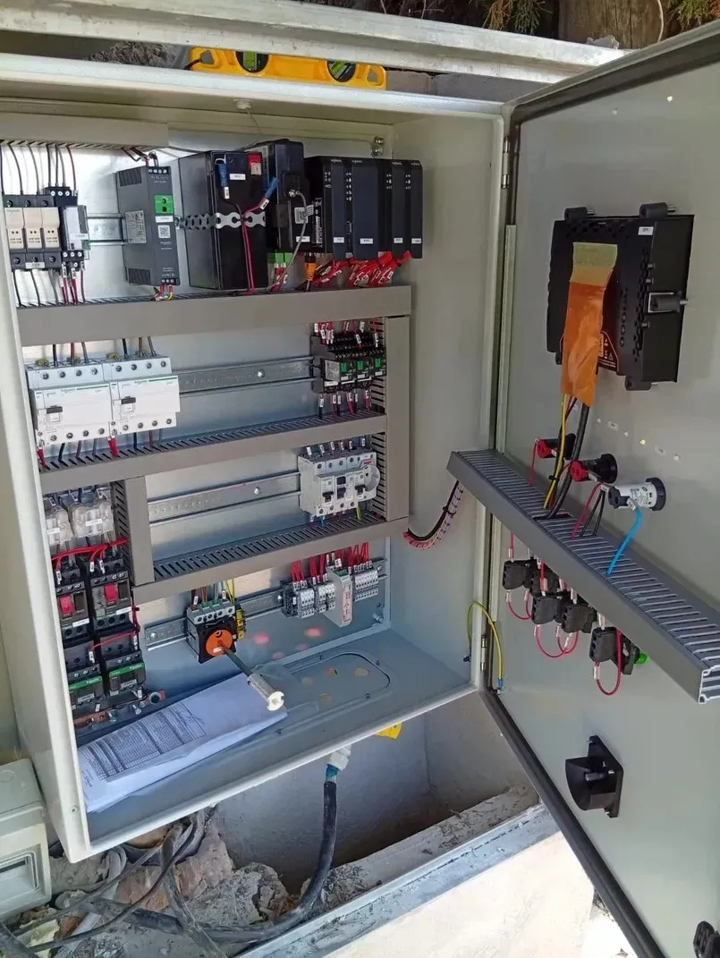 Panel de control con varios componentes eléctricos, cables y un documento dentro de una carcasa metálica abierta.