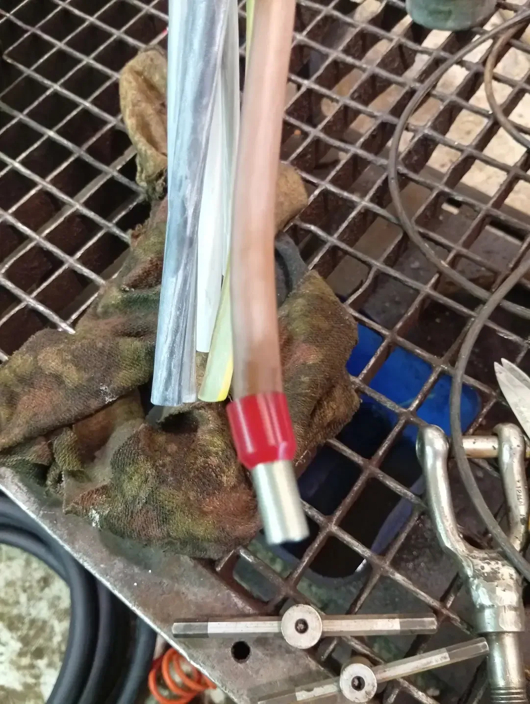 Primer plano de un cable cortado con el extremo rojo aislado sobre una rejilla metálica. También se ven varias herramientas y un guante.
