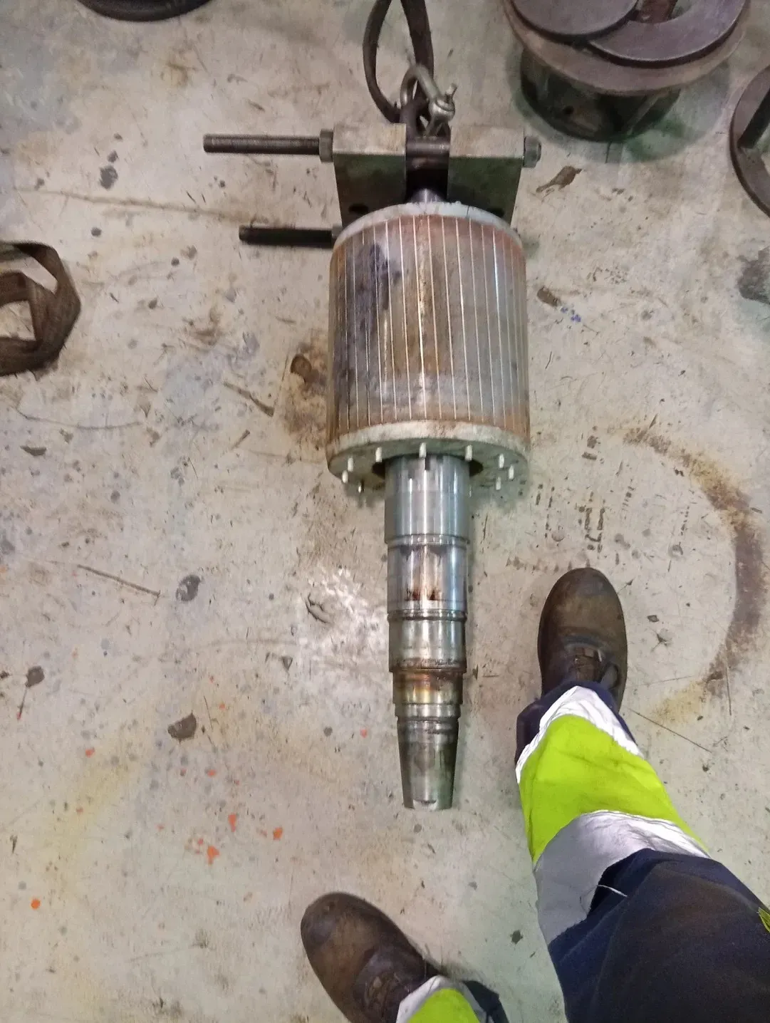 Un objeto metálico cilíndrico, probablemente un rotor, suspendido sobre el suelo. Las piernas y los pies de una persona aparecen en el marco.