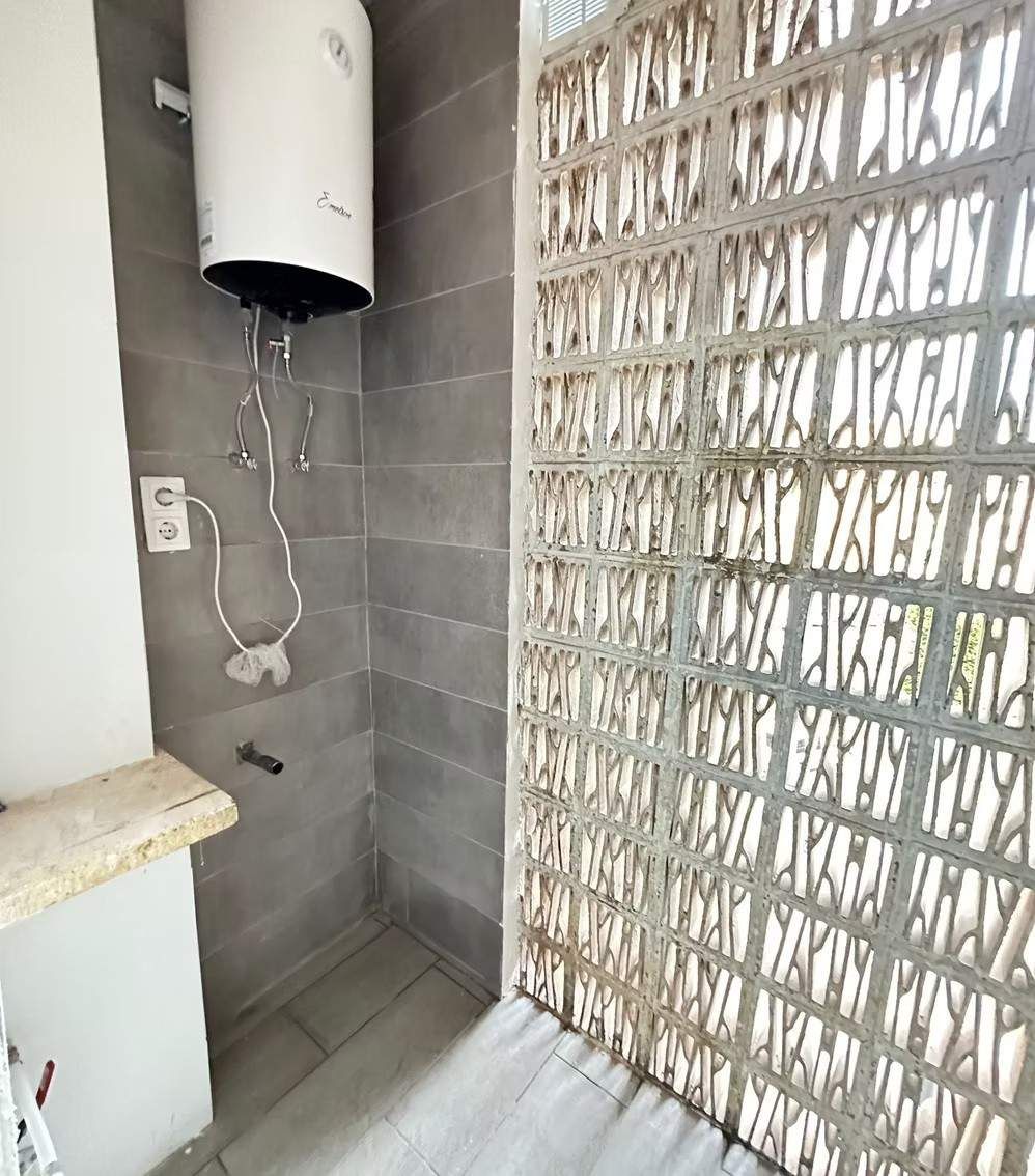 Pequeño baño con calentador de agua en la pared, azulejos grises y mampara de ladrillo blanco estampado.