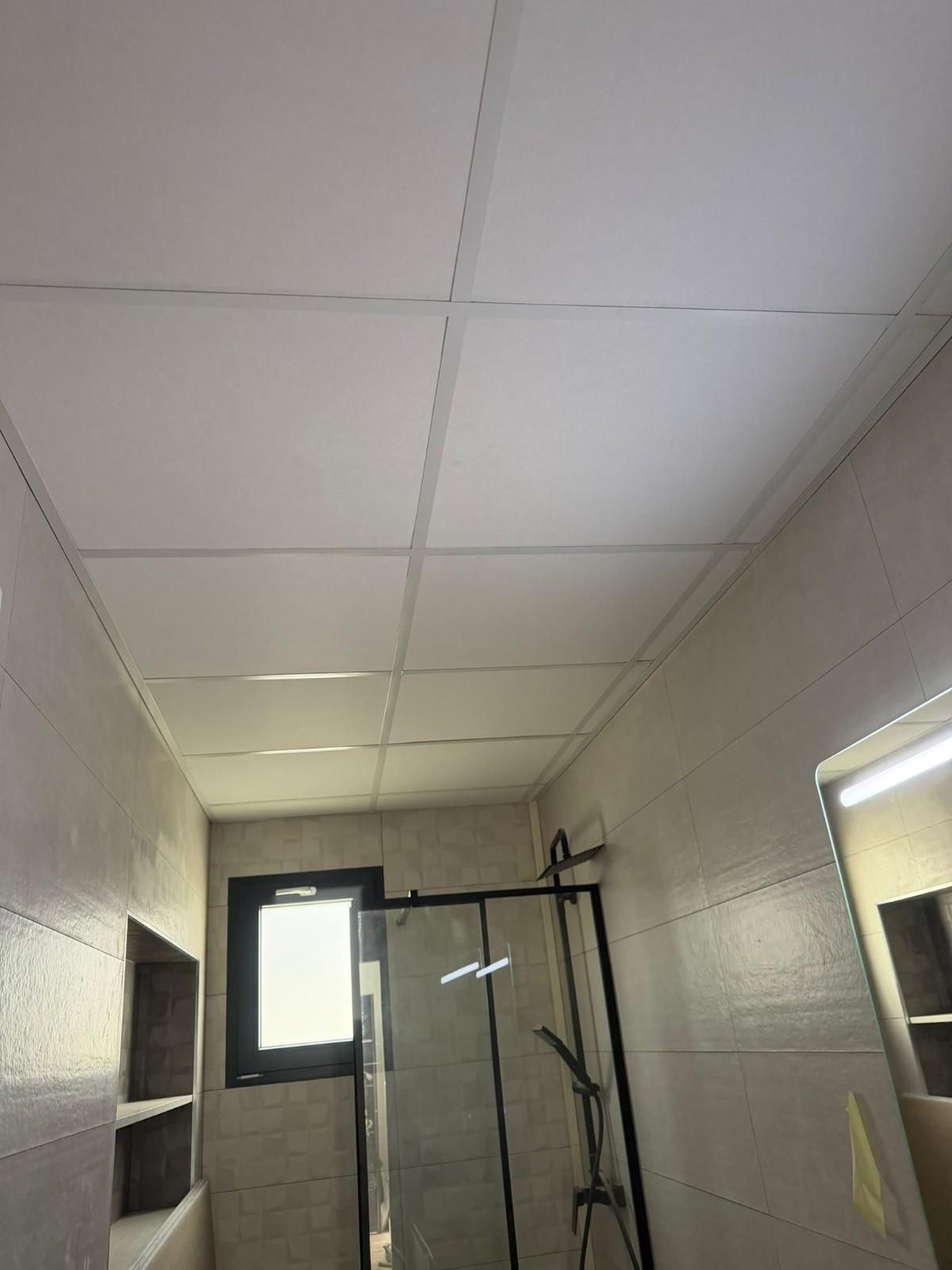 Baño con techo y paredes de azulejos blancos, ducha con puerta de cristal y pequeña ventana.