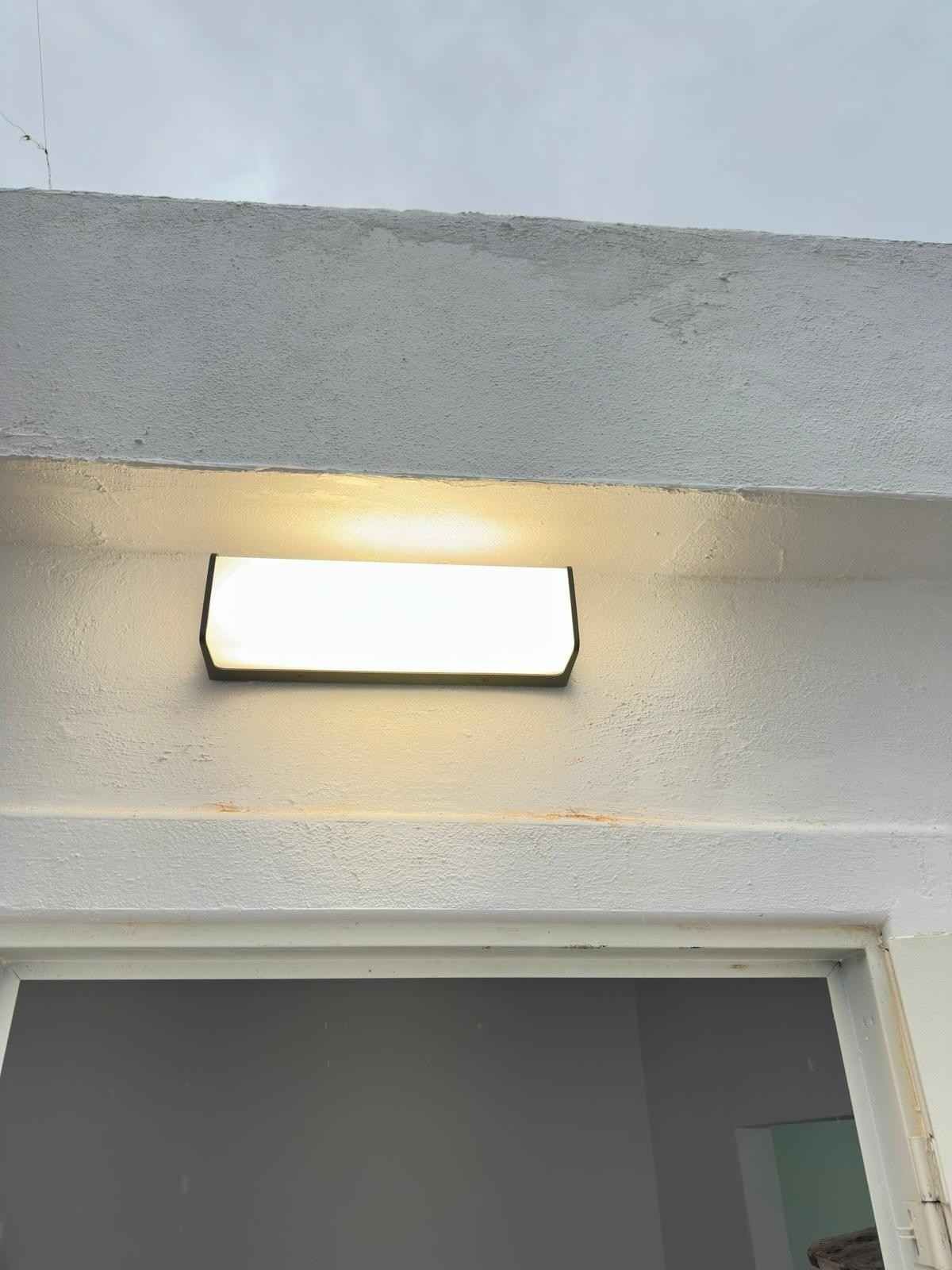 Lámpara rectangular blanca montada encima de una puerta, iluminada contra una pared gris.