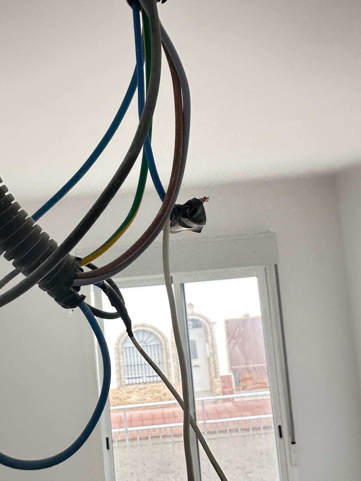 Cables eléctricos y conductos expuestos colgando de un techo blanco, cerca de una ventana.