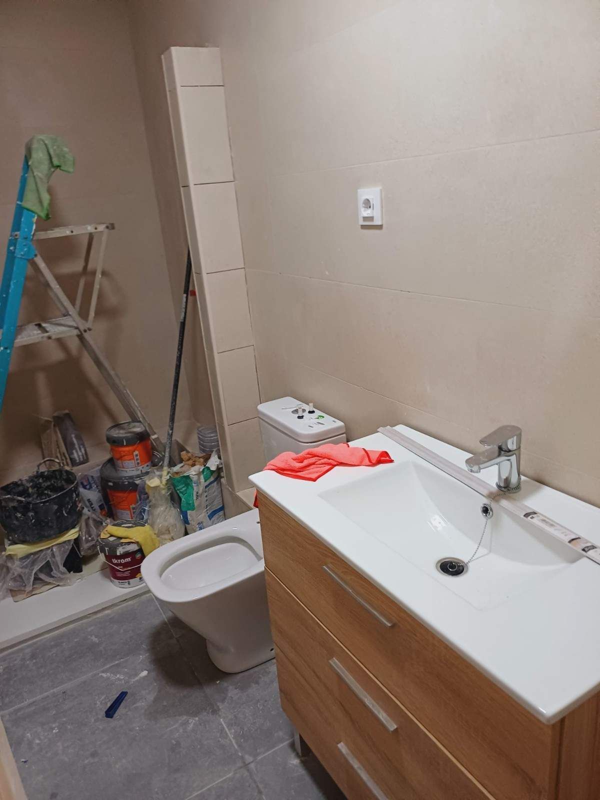 Baño en reformas, con inodoro, lavabo, armario, armario alto, escalera y herramientas. Paredes beige, suelo gris.