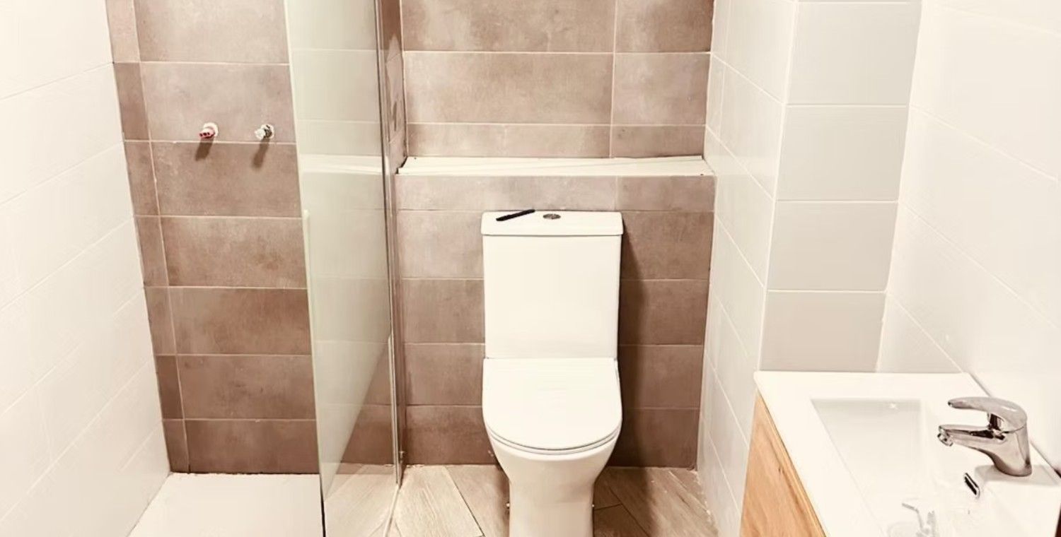 Baño con inodoro centrado contra la pared de azulejos. Ducha a la izquierda, lavabo a la derecha. Azulejos beige y blancos.