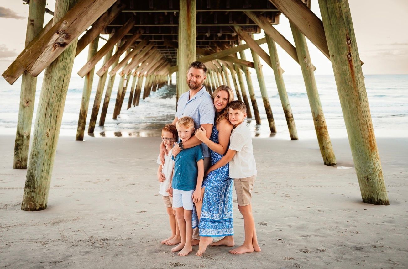 A family of five posing under a wooden pier on a sandy beach at sunset.