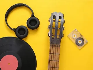 Auriculares negros, mástil de guitarra, disco de vinilo y cinta de casete sobre fondo amarillo.