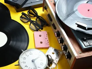 Discos de vinilo, tocadiscos, cintas de casete y vasos sobre superficie amarilla.