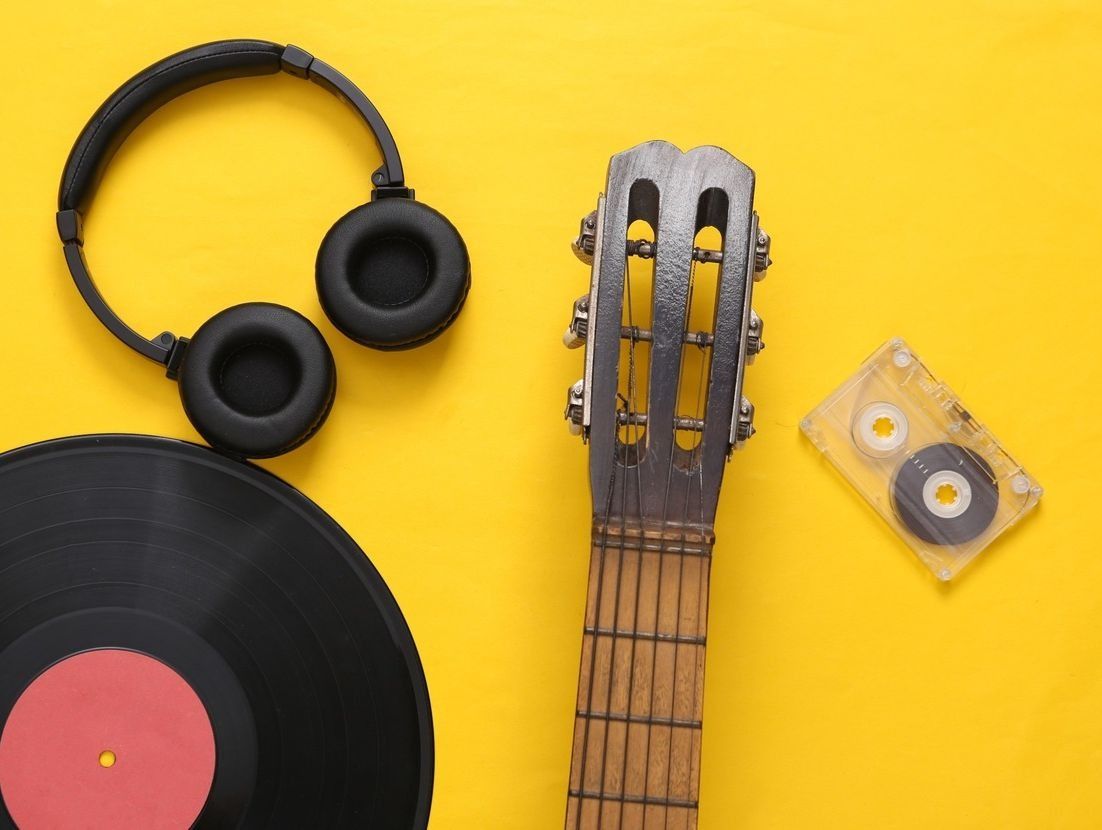 Disco de vinilo negro, auriculares, mástil de guitarra y cinta de casete sobre fondo amarillo.