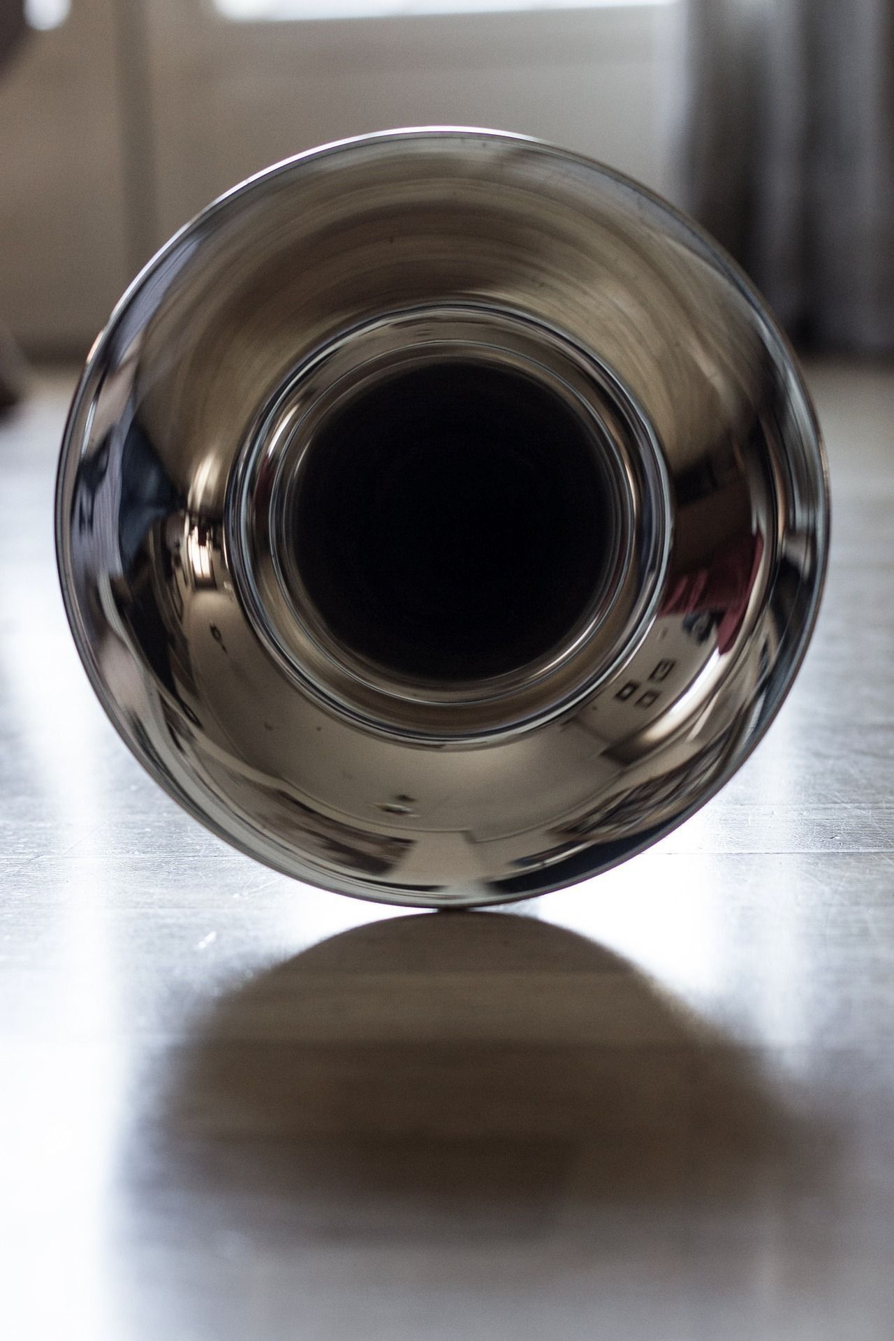 Tubo de escape de metal brillante, abertura interior de color negro, que reposa sobre un suelo de madera, proyectando una sombra.