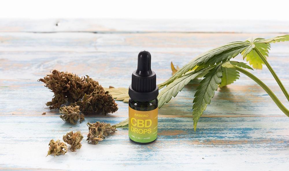 Una botella de aceite de CBD con gotero, rodeada de cogollos de cannabis secos y hojas verdes sobre un fondo rústico de madera azul.