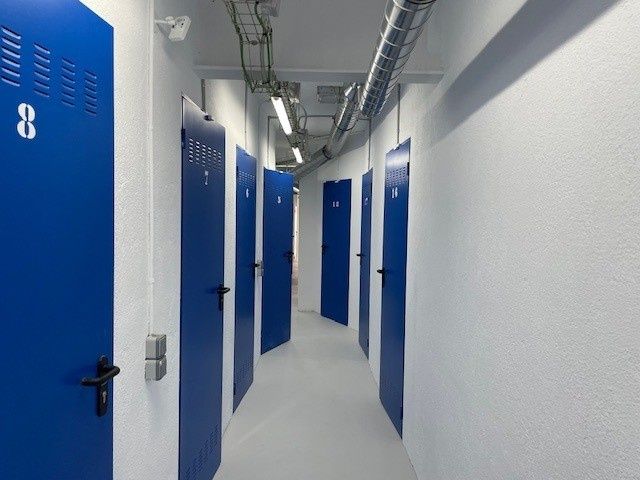 Taquillas azules bordean un pasillo blanco impecable con tuberías y rejillas de ventilación en el techo.