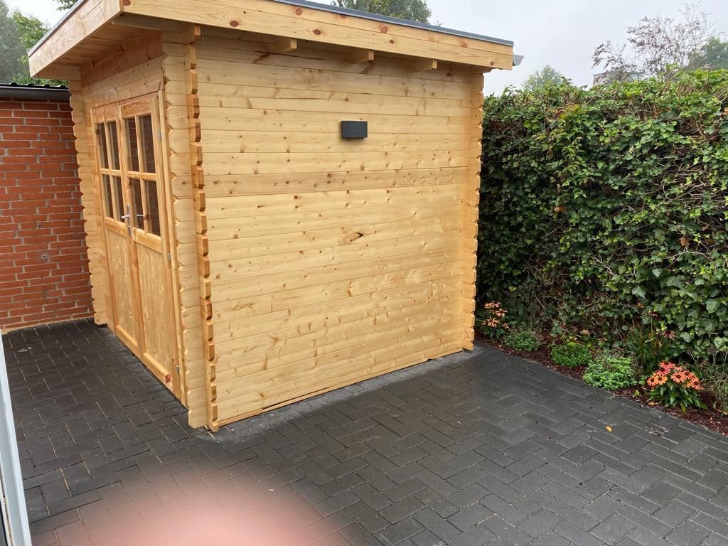 Auf einer gemauerten Terrasse neben einer Hecke steht ein kleiner Holzschuppen.