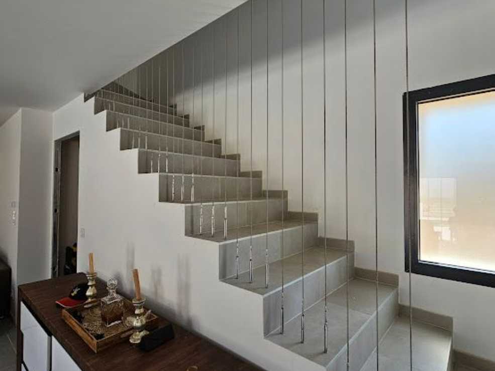 Escalier aux marches grises, murs blancs et rampe moderne en câbles. On aperçoit une fenêtre à cadre sombre.