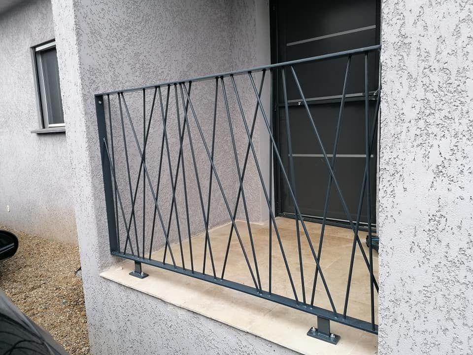 Garde-corps de balcon en métal gris avec barreaux diagonaux, sur un bâtiment aux murs gris texturés.
