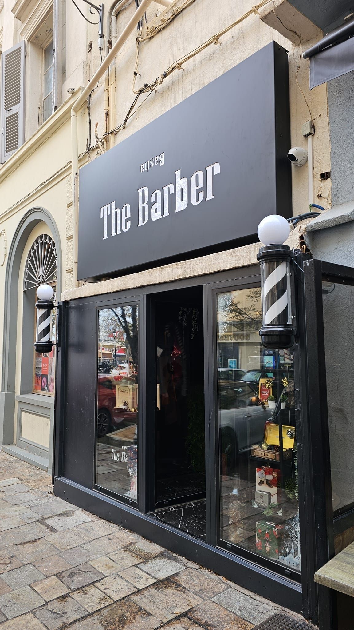Panneau de salon de coiffure noir et blanc indiquant The Barber au-dessus d'une porte vitrée, avec poteau de barbier.