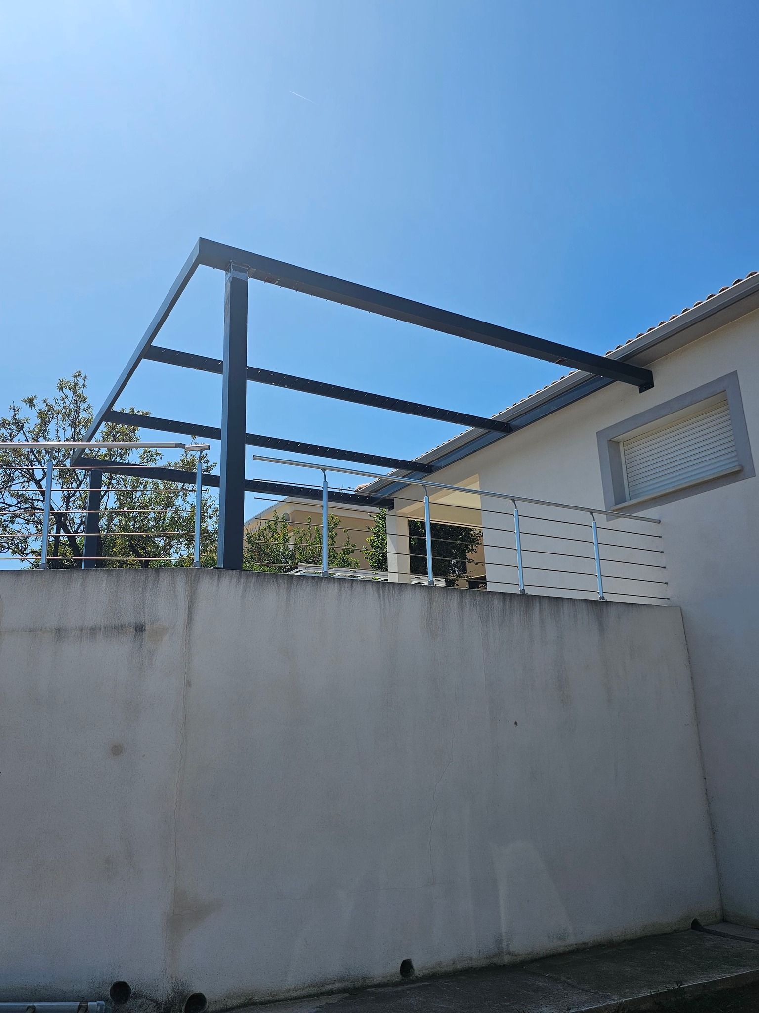 Pergola en métal noir sur un mur blanc, sous un ciel bleu clair.