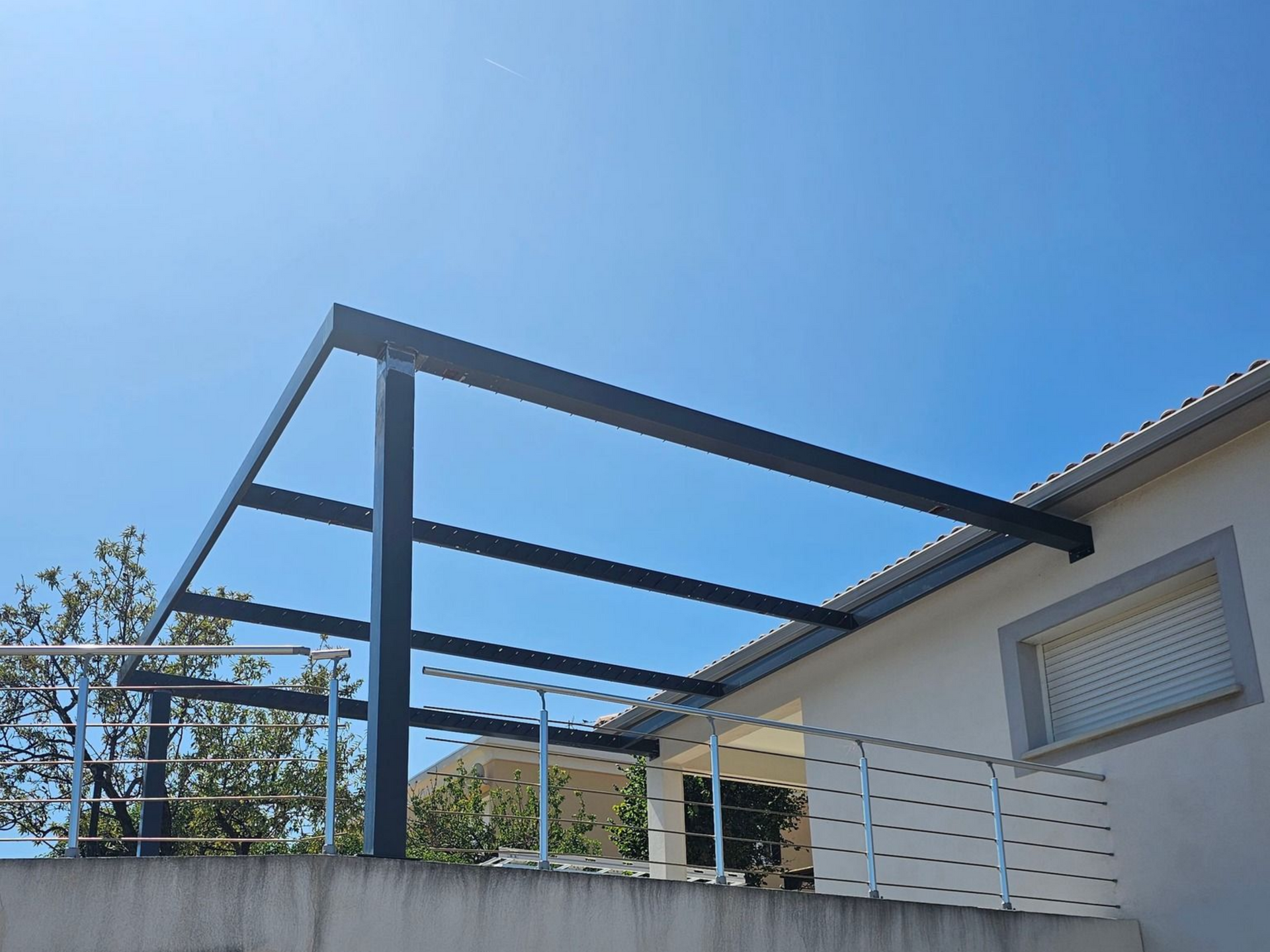 Pergola en métal noir sur un bâtiment blanc contre un ciel bleu clair.