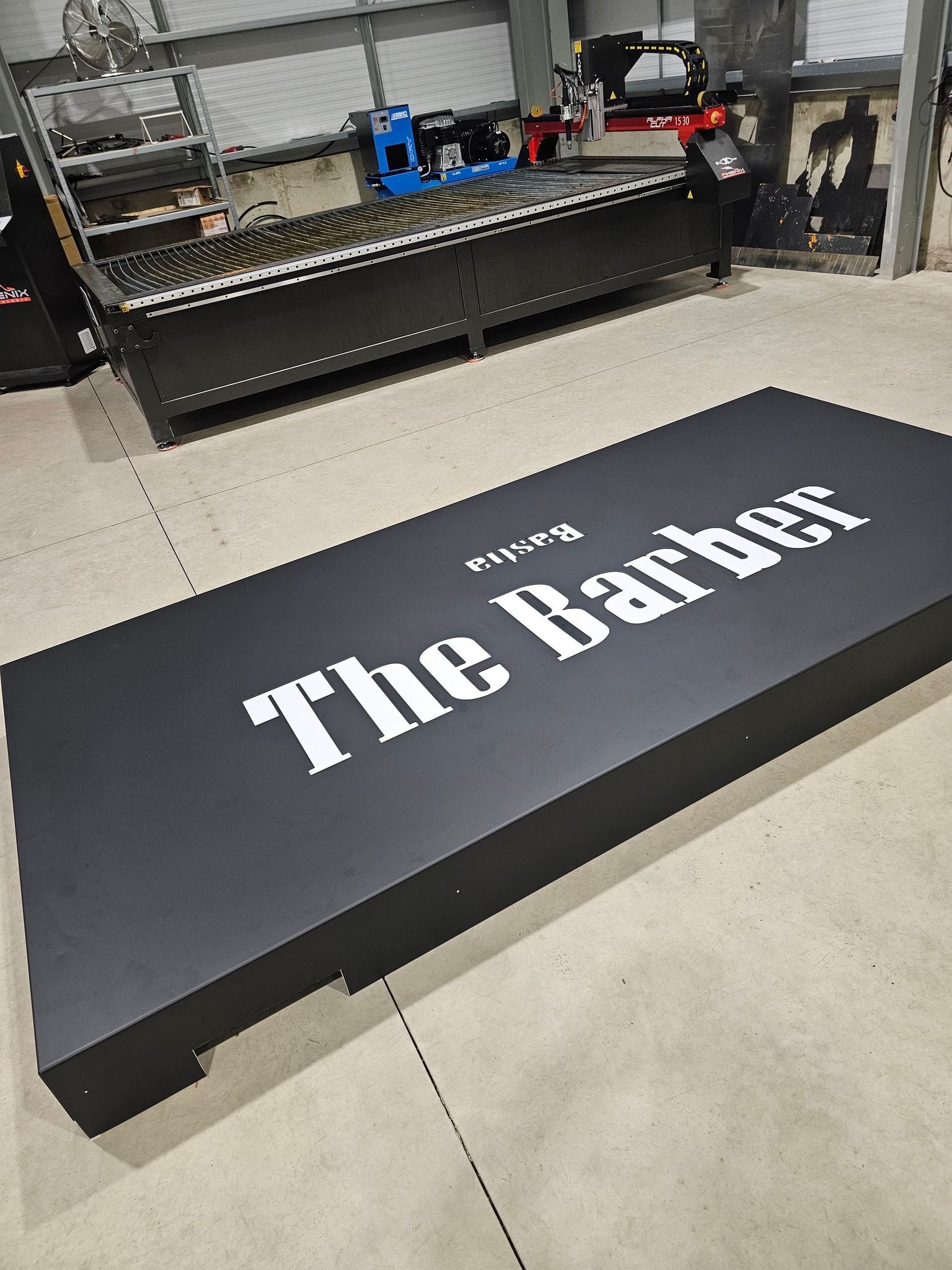 Panneau noir d'une enseigne sur lequel est écrit The Barber en blanc, près d'une grande machine dans un atelier.