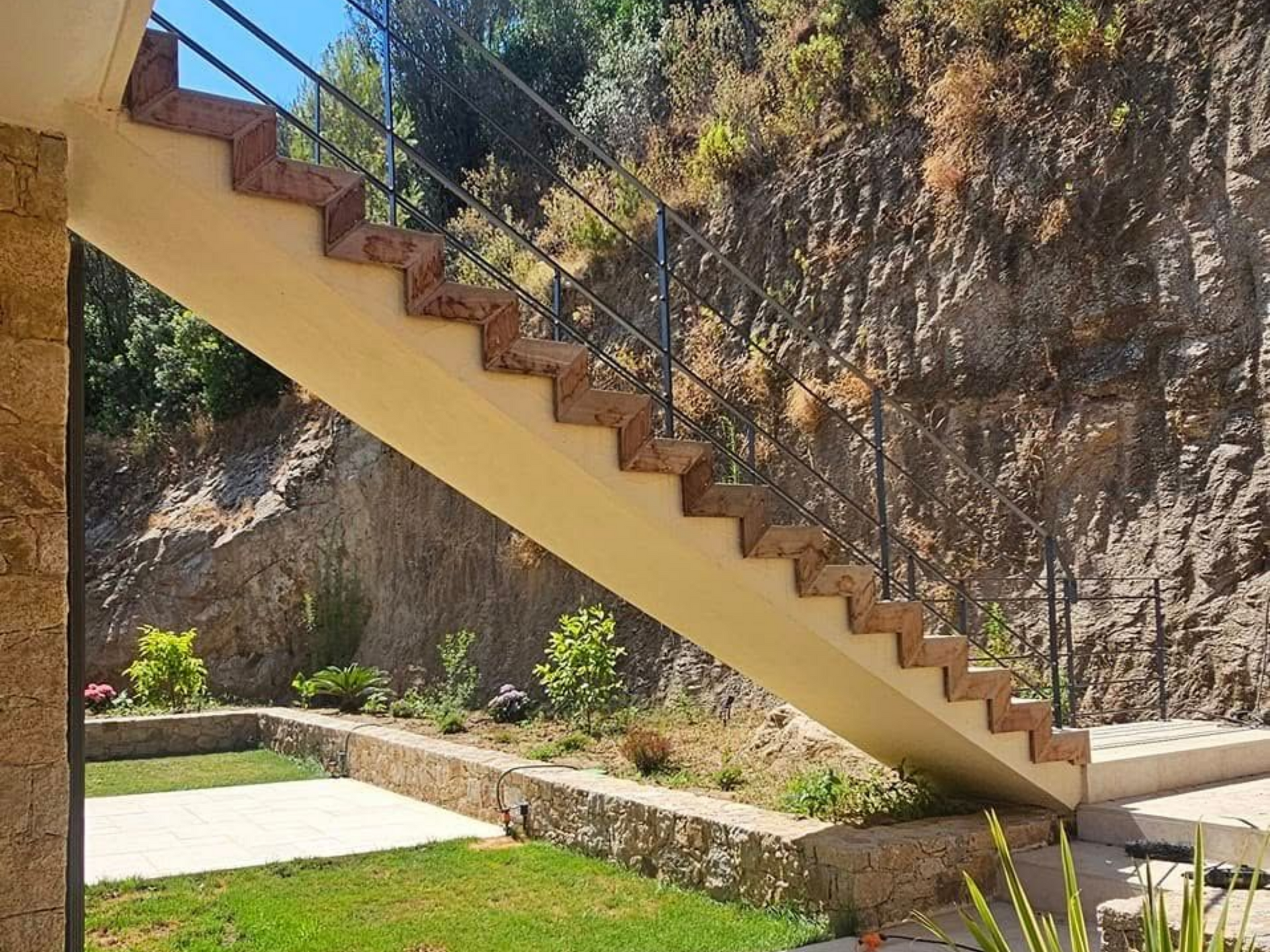 Un escalier extérieur avec des marches rouge brun et une rampe en métal monte une colline.